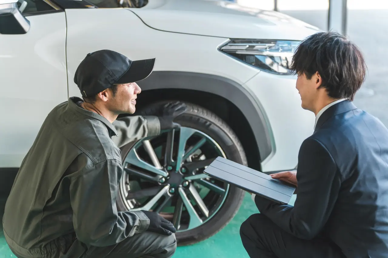 整備士が、車のタイヤを指差しながら、スーツ姿の男性と見積もりやプランについて相談している様子